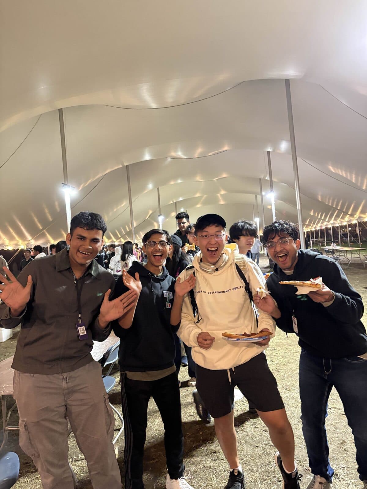 Four friends laughing over pizza under an event tent at night